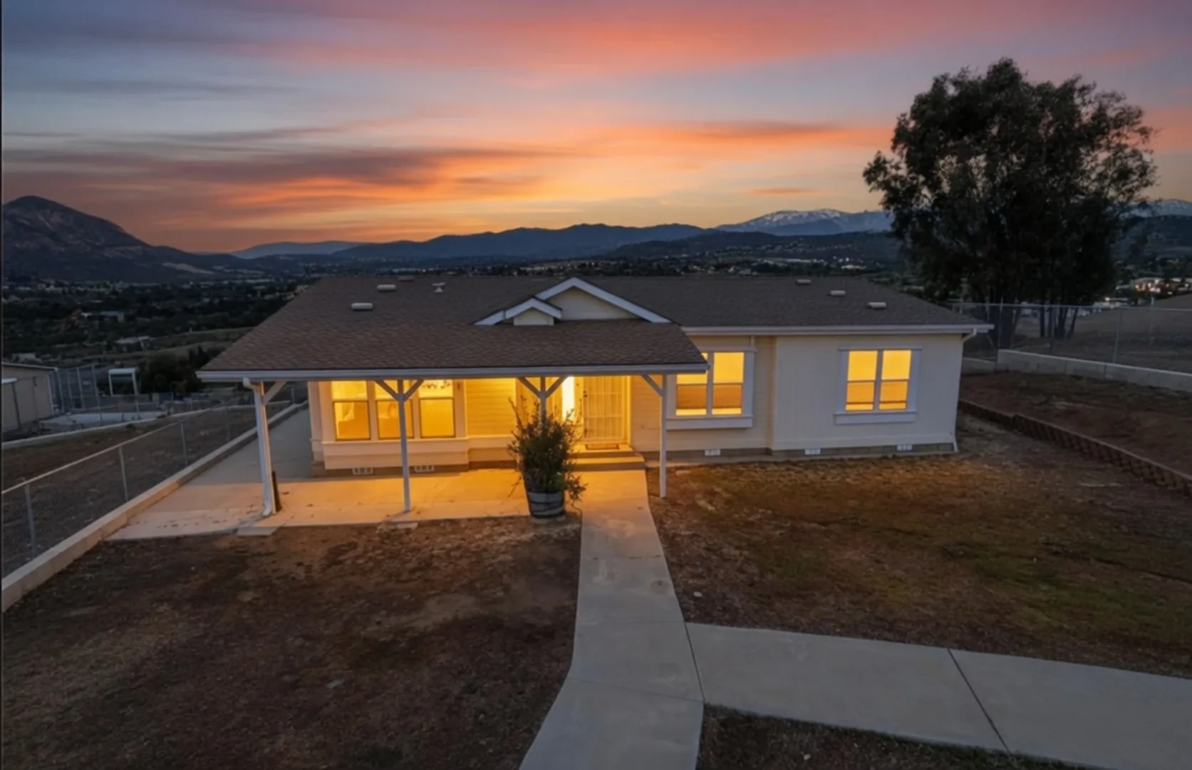 Front of 39210 Grassy Rd at sunset, single-story home with covered porch lit warmly against a pink and orange sky over the foothills.